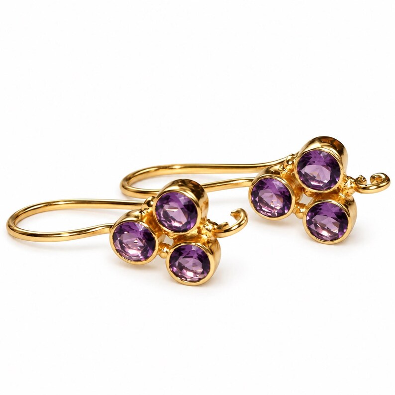 May include: A pair of gold earrings featuring three round, faceted purple amethyst gemstones arranged in a cluster. Each earring is attached to a curved gold hook wire, showcasing a polished metallic finish and vibrant violet stones.