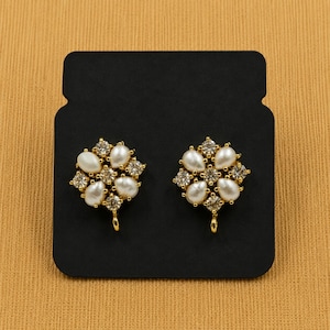 May include: A pair of gold-toned stud earrings. Each earring features a floral design with white pear-shaped faux pearls and clear, square-cut crystals. The earrings are displayed on a black card.