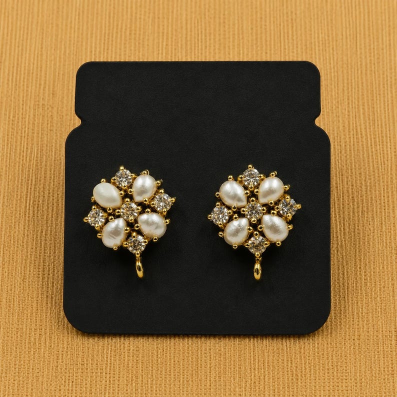 May include: A pair of gold-toned stud earrings. Each earring features a floral design with white pear-shaped faux pearls and clear, square-cut crystals. The earrings are displayed on a black card.