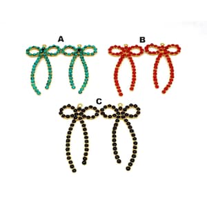 May include: Three pairs of gold-tone bow-shaped charms with crystal embellishments. The bows are adorned with turquoise, red, and black crystals.