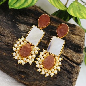 May include: A pair of gold tone earrings with brown and white stone accents. The earrings feature a square shaped stone, a teardrop shaped stone, and a cluster of white pearls.
