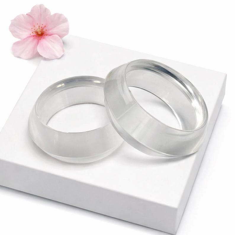 May include: Two clear, oval-shaped bracelets are displayed on a white surface, with a pink flower in the background. The bracelets are made of a transparent material, reflecting light. The overall aesthetic is minimalist and elegant.