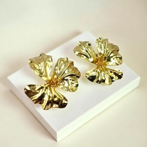 Może przedstawiać: Dwie złote kolczyki w kształcie kwiatów. Każdy kolczyk ma pięć płatków i teksturowaną powierzchnię. Kolczyki są prezentowane na białej powierzchni na białym tle. Kolczyki są wyrazistym elementem biżuterii.