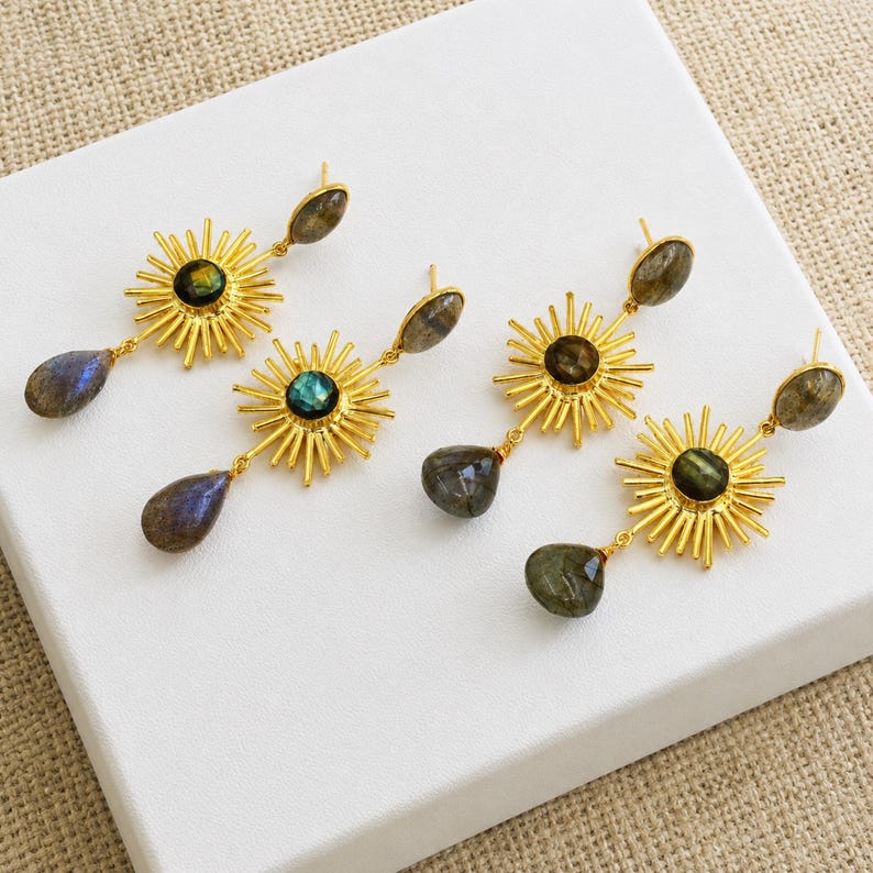 Puede incluir: Dos pares de pendientes dorados con un dise&ntilde;o de rayos de sol. Cada pendiente tiene una piedra de labradorita redonda e iridiscente en el centro y una piedra de labradorita en forma de l&aacute;grima que cuelga de la parte inferior del sol.