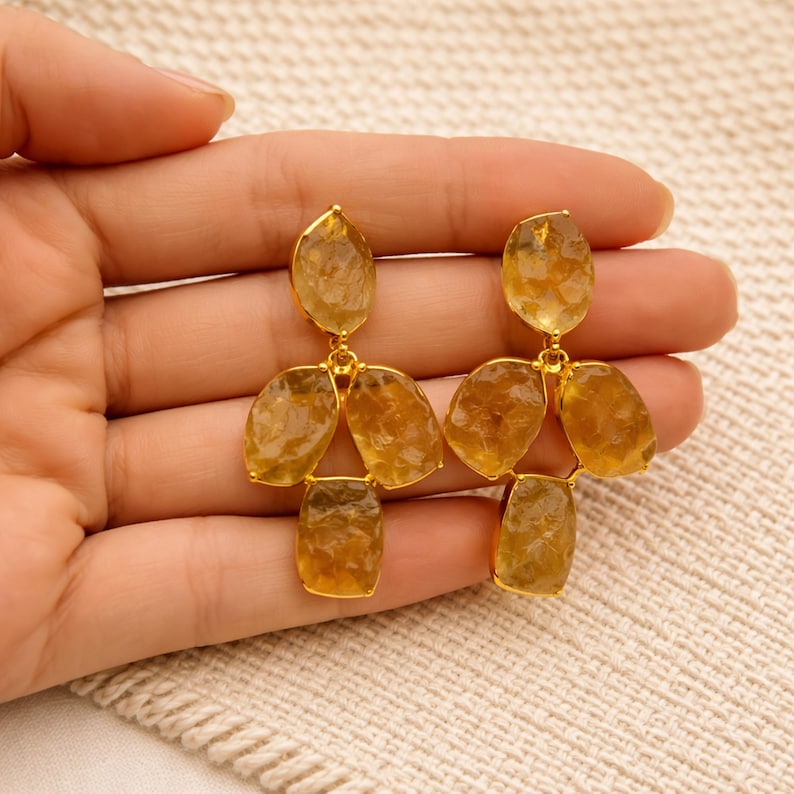 May include: Gold-toned earrings featuring clusters of faceted, amber-colored gemstones. The earrings have a three-dimensional design, with each earring composed of multiple stones connected by delicate gold links. The gemstones have a natural, irregular shape.