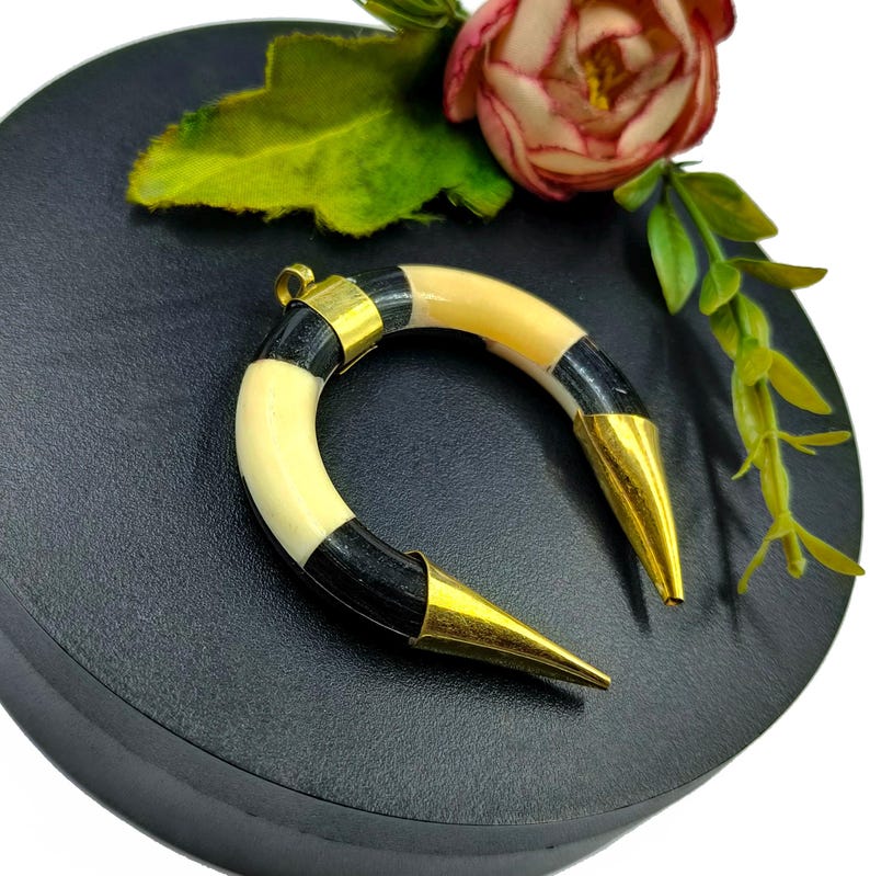 Peut inclure: Un pendentif en forme de croissant compos&eacute; de segments altern&eacute;s de mati&egrave;re lisse noire et cr&egrave;me. La pi&egrave;ce est agr&eacute;ment&eacute;e d'embouts en m&eacute;tal dor&eacute; poli aux deux extr&eacute;mit&eacute;s pointues et d'une b&eacute;li&egrave;re centrale en m&eacute;tal dor&eacute; pour la suspension.