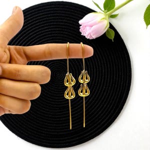 May include: A pair of gold-colored drop earrings. Each earring features a stylized floral design with three petal-like shapes stacked on a long, slender post. The earrings are displayed against a black textured background, with a pink rose in the background.