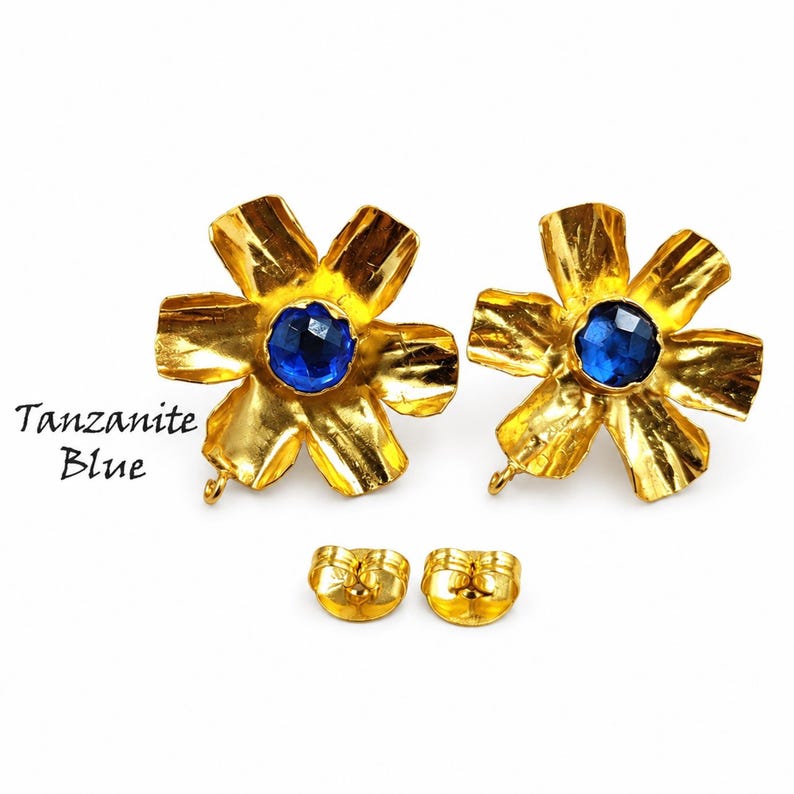 May include: A pair of gold-toned flower-shaped stud earrings with a textured finish and a faceted tanzanite blue center stone. The earrings include two matching gold-toned earring backs. The text Tanzanite Blue is visible on the left side.
