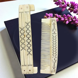 Puede incluir: Dos peines antiguos de marfil. Uno es un peine plegable con un diseño geométrico decorativo en tinta negra. El otro es un peine estándar con un diseño similar. Ambos peines se exhiben sobre una superficie azul oscuro con flores moradas.