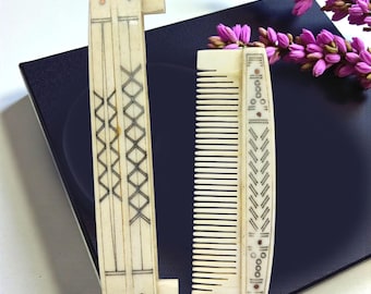 May include: Two antique ivory combs. One is a folding comb with a decorative geometric design in black ink. The other is a standard comb with a similar design. Both combs are displayed on a dark blue surface with purple flowers.