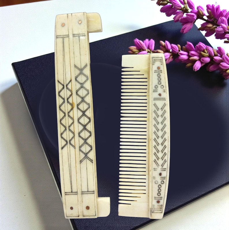 May include: Two antique ivory combs. One is a folding comb with a decorative geometric design in black ink. The other is a standard comb with a similar design. Both combs are displayed on a dark blue surface with purple flowers.