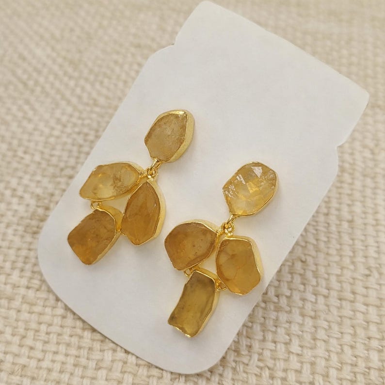 May include: Gold-toned dangle earrings featuring clusters of rough-cut, amber-colored gemstones. The earrings are displayed on a white card against a neutral background. The gemstones have a natural, irregular shape.