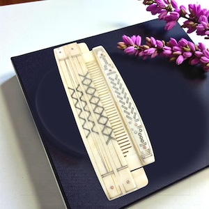 May include: Two antique ivory-colored combs with intricate geometric designs. One comb features a series of intersecting lines, while the other has a pattern of diagonal lines and circles. The combs are displayed on a dark blue surface, with purple flowers in the background.