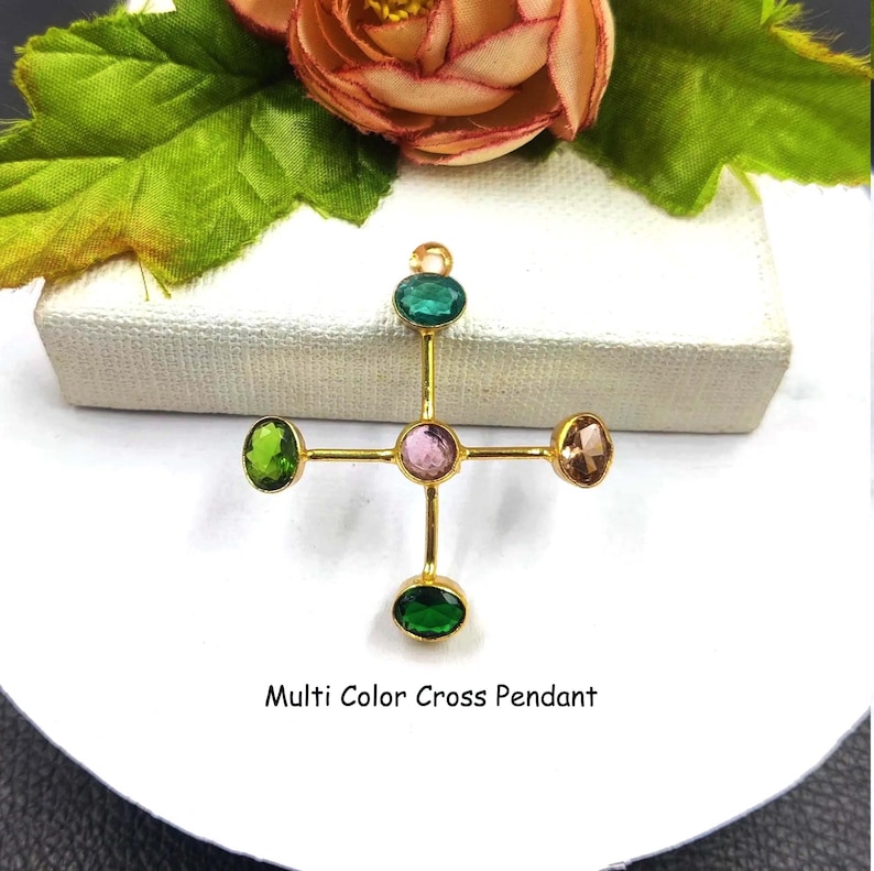 Puede incluir: Un colgante de cruz dorada con cinco piedras preciosas de colores. Las piedras preciosas son verdes, rosas y marrones. El texto "Multi Color Cross Pendant" est&aacute; debajo.