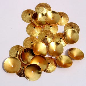 May include: A group of 17 gold-colored metal embellishments with a textured surface. The embellishments are round and have a small hole in the center. They are arranged in a loose pile on a white surface.