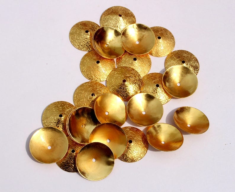 May include: A group of 17 gold-colored metal embellishments with a textured surface. The embellishments are round and have a small hole in the center. They are arranged in a loose pile on a white surface.