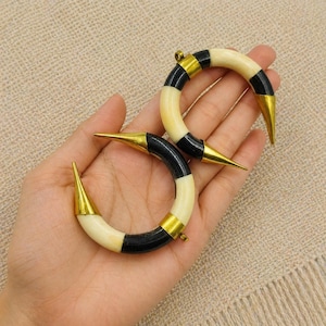 Peut inclure: Deux boucles d'oreilles cr&eacute;oles en forme de croissant compos&eacute;es de segments altern&eacute;s en mat&eacute;riau cr&egrave;me lisse et en mat&eacute;riau noir poli, orn&eacute;es d'embouts m&eacute;talliques dor&eacute;s pointus &agrave; chaque extr&eacute;mit&eacute; de la structure courbe.