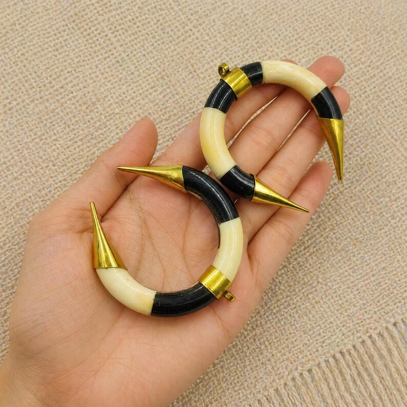 Peut inclure: Deux boucles d'oreilles cr&eacute;oles en forme de croissant compos&eacute;es de segments altern&eacute;s en mat&eacute;riau cr&egrave;me lisse et en mat&eacute;riau noir poli, orn&eacute;es d'embouts m&eacute;talliques dor&eacute;s pointus &agrave; chaque extr&eacute;mit&eacute; de la structure courbe.