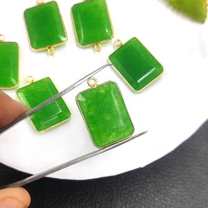 Puede incluir: Se muestran varios colgantes rectangulares de jade verde con marcos dorados. Un colgante está siendo sujetado por unas pinzas. Los colgantes tienen un lazo en la parte superior para sujetarlos a una cadena u otro componente de joyería.