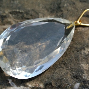 May include: A clear, faceted crystal pendant with a teardrop shape. The crystal has a gold-colored bail for hanging. The pendant is displayed on a dark, textured surface, highlighting its clarity and cut.