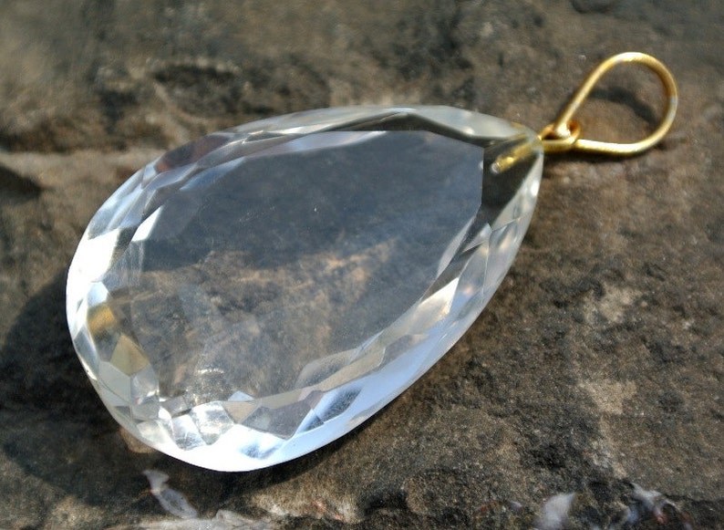 May include: A clear, faceted crystal pendant with a teardrop shape. The crystal has a gold-colored bail for hanging. The pendant is displayed on a dark, textured surface, highlighting its clarity and cut.