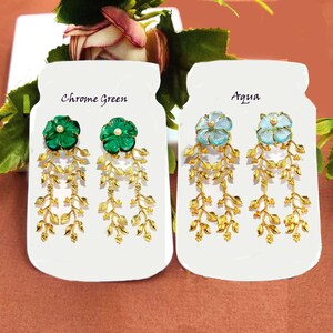 Puede incluir: Dos pares de pendientes colgantes con dise&ntilde;os de hojas de oro y piedras de vidrio en forma de flor. Los pendientes se muestran en tarjetas blancas con las palabras "Chrome Green" y "Aqua" impresas en ellas.