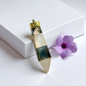 Puede incluir: Un colgante geométrico con una tapa dorada y un diseño en capas de materiales negros, blancos e iridiscentes. El colgante está sobre un fondo blanco con una flor morada.