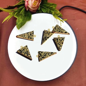 Puede incluir: Seis colgantes triangulares de jaspe dálmata con anillas de salto doradas. Los colgantes están en un plato blanco.