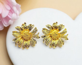 May include: A pair of gold-coloured daisy stud earrings. The earrings feature textured petals and a textured centre, resembling a real daisy. The earrings are displayed on a white heart-shaped surface.