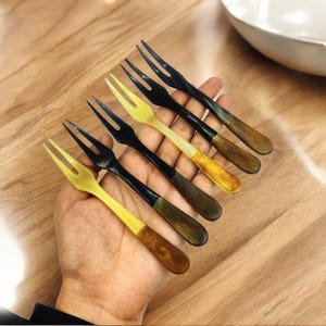 May include: A set of six small forks with two tines each. The forks have black tines and handles that transition from yellow to brown. The forks are held in a hand, with a wooden surface in the background.