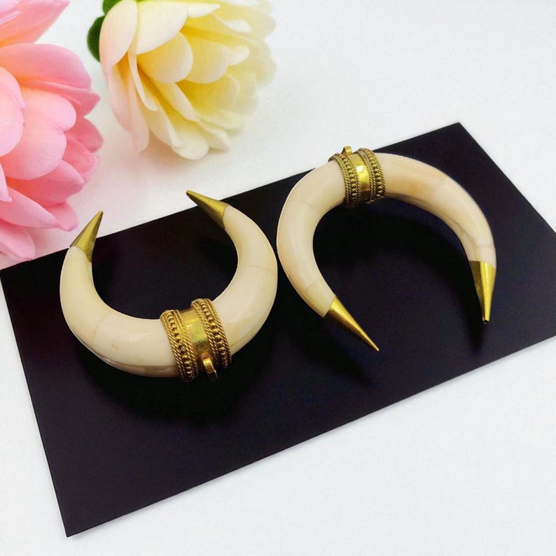 May include: Two ivory-coloured, crescent-shaped earrings with gold-coloured tips and decorative bands. The earrings are displayed on a black surface, with pink and yellow flowers in the background. The earrings have a bohemian or tribal aesthetic.