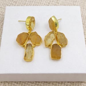 May include: A pair of gold-toned earrings featuring raw, amber-colored gemstones. Each earring has a post and a cluster of four stones in varying shapes, creating a unique, natural look. The earrings are displayed on a white surface.