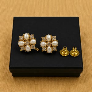 May include: A pair of gold-toned, floral-shaped earrings with white pearl and clear crystal accents, presented on a black box. The earrings have a post and backing. The earrings are a jewelry item.