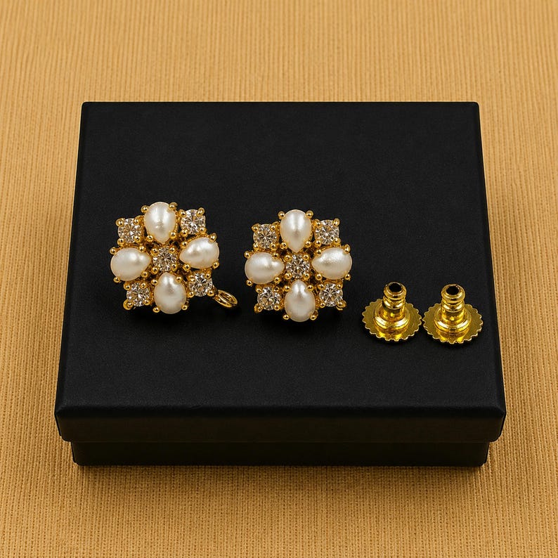 May include: A pair of gold-toned, floral-shaped earrings with white pearl and clear crystal accents, presented on a black box. The earrings have a post and backing. The earrings are a jewelry item.