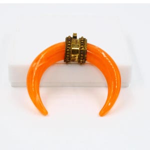 May include: Orange crescent moon pendant with a gold-toned metal accent. The pendant is made of glass and has a smooth, polished finish.