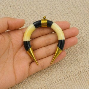 Peut inclure: Un pendentif en forme de croissant compos&eacute; de segments altern&eacute;s de couleur cr&egrave;me et noire. La pi&egrave;ce est agr&eacute;ment&eacute;e d'un connecteur central en m&eacute;tal dor&eacute; sur le dessus et d'embouts pointus en m&eacute;tal dor&eacute; aux deux extr&eacute;mit&eacute;s de la courbe.