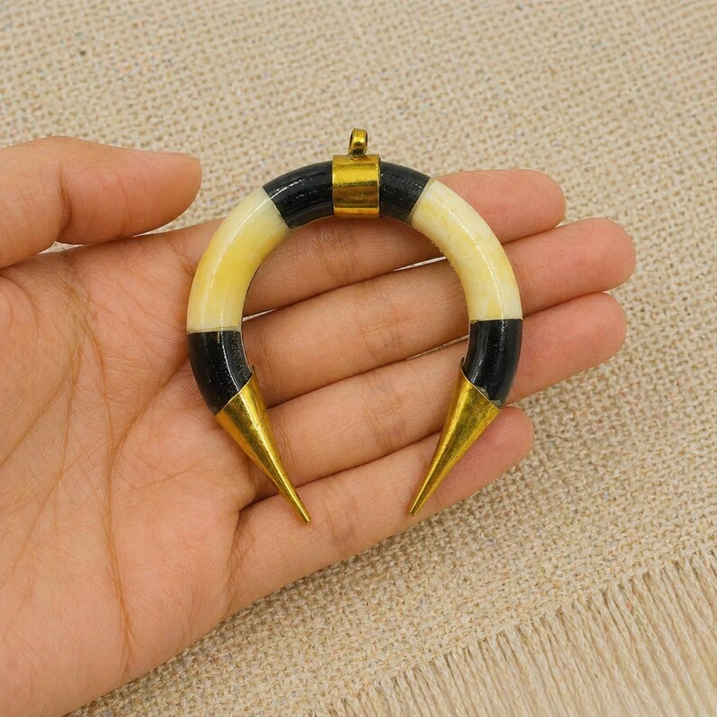Peut inclure: Un pendentif en forme de croissant compos&eacute; de segments altern&eacute;s de couleur cr&egrave;me et noire. La pi&egrave;ce est agr&eacute;ment&eacute;e d'un connecteur central en m&eacute;tal dor&eacute; sur le dessus et d'embouts pointus en m&eacute;tal dor&eacute; aux deux extr&eacute;mit&eacute;s de la courbe.
