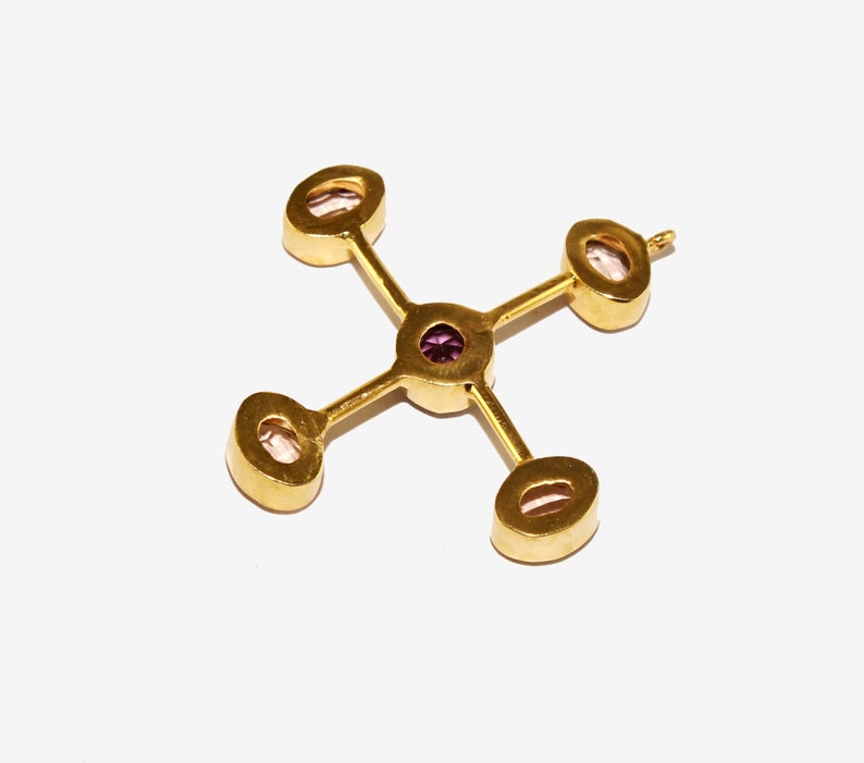 Puede incluir: Colgante dorado con dise&ntilde;o de cruz. Cuenta con cuatro piedras ovaladas de color claro en los extremos y una peque&ntilde;a piedra redonda morada en el centro. El colgante se encuentra sobre un fondo blanco.
