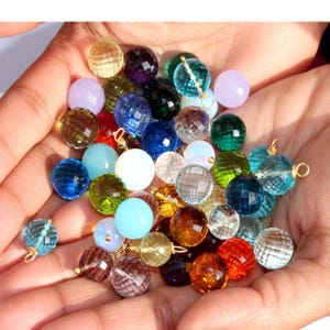 May include: A collection of faceted gemstone beads in various colors, including clear, blue, green, purple, and orange. Each bead has a gold loop attached for jewelry making.