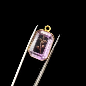 May include: A rectangular pendant featuring a faceted light purple stone set in a gold-toned metal frame. Inside the transparent stone, a small brown twig with several round seed pods is visible. A small gold-toned loop is attached to the top.