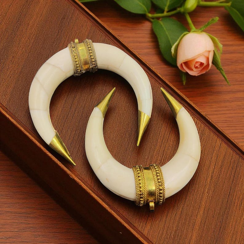 May include: Two ivory-coloured, crescent-shaped pendants with gold-coloured metal accents. The pendants have pointed tips and are adorned with decorative bands of gold-coloured metal. The pendants are displayed on a wooden surface.