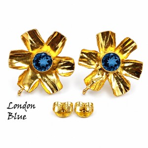 May include: A pair of gold-toned flower-shaped earrings featuring a central round faceted London Blue gemstone. Each earring has six textured petals and a small loop at the base. Two matching gold-toned earring backs are included below the earrings.