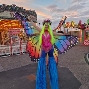 Może przedstawiać: Osoba w kostiumie motyla z tęczowymi skrzydłami, różowym topem z cekinami, niebieskimi spodniami i niebieskimi osłonami na szczudła. Osoba ma na sobie wianek z kwiatów i perukę w kolorze neonowej zieleni. W tle widać karuzele.