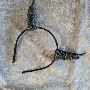 Könnte beinhalten: Ein schwarzes Stirnband mit zwei schwarzen und weißen Hörnern. Das Stirnband ist aus weichem, flexiblem Material und die Hörner aus hartem, strukturiertem Material.