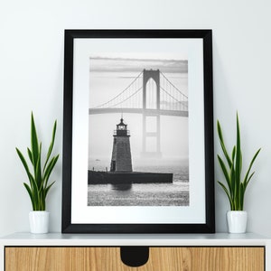 May include: A black and white photograph of a lighthouse and a bridge in the distance. The lighthouse is in the foreground and the bridge is in the background. The photo is taken on a foggy day.