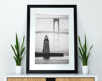 May include: A black and white photograph of a lighthouse and a bridge in the distance. The lighthouse is in the foreground and the bridge is in the background. The photo is taken on a foggy day.