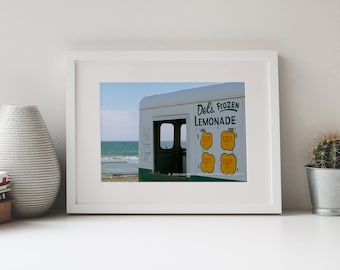 May include: A white vintage ice cream truck with green trim parked on a sandy beach. The truck has a sign that reads "Del's Frozen Lemonade" with prices listed for different sizes. The ocean is visible in the background.