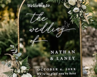 Può includere: Un cartello nuziale a specchio ad arco con cornice dorata, decorato con rose bianche e felci verdi. Il testo recita: Welcome to the wedding of NATHAN & LANEY, OCTOBER 4, 20XX, We're so glad you're here. Il cartello presenta eleganti scritte bianche.