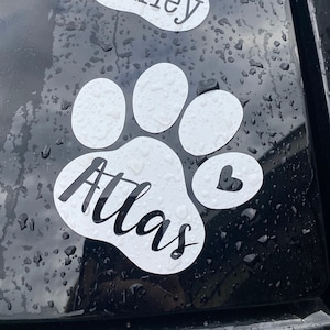 May include: Two white paw print decals with the names "Oakley" and "Atlas" written in black cursive font. Each paw print has a small heart in the top right corner.