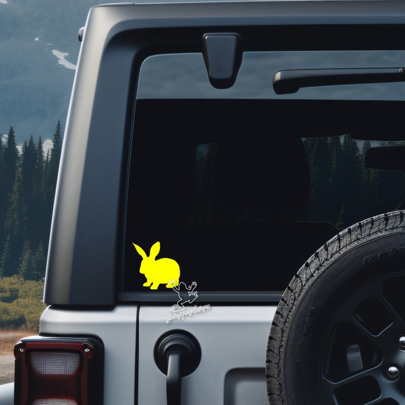 Peut inclure: Une silhouette jaune de lapin avec une silhouette blanche de grenouille derri&egrave;re elle. L'autocollant est sur la vitre arri&egrave;re d'un SUV gris. Le texte "Hoppy Days" est sous l'autocollant.
